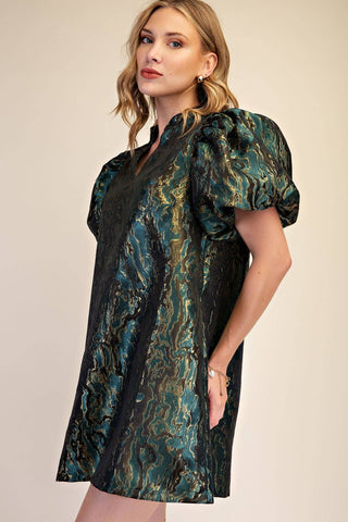 A woman modeling a short puff-sleeve dress with a black base and an abstract teal, green, and gold marbled pattern, set against a plain beige studio background.