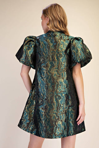 A woman shown from the back wearing a short puff-sleeve dress with a shimmering teal, green, and gold marbled pattern on a black base, set against a plain beige studio background.