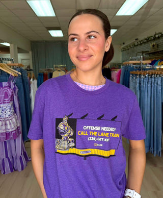 A woman wearing a purple graphic t-shirt with a cartoon train and the text 'OFFENSE NEEDED! CALL THE LANE TRAIN (225) GET-OUT' in a clothing store.