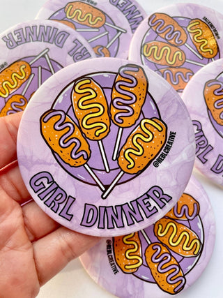 Sticker with corn dogs and 'Girl Dinner' text on a purple background, held by a hand.