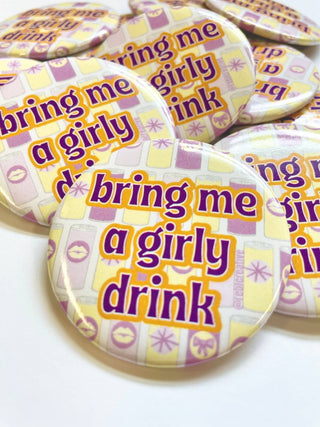 A close-up of round pin-back buttons featuring the phrase 'bring me a girly drink' in purple text with a yellow outline, set against a pastel pattern of yellow, lavender, and white.