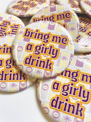 A pile of round pin-back buttons featuring the phrase 'bring me a girly drink' in purple text with an orange outline, set against a patterned background of pastel yellow, lavender, and white.