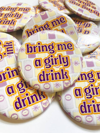 A pile of round pin-back buttons featuring the phrase 'bring me a girly drink' in purple text with a yellow outline, set against a pale yellow and lavender patterned background.