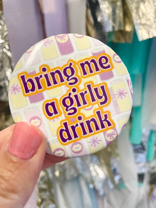 A hand holding a round pin-back button with the phrase 'bring me a girly drink' in purple text outlined in yellow, set against a patterned background of pastel pink, yellow, and white.