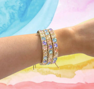 A close-up of a person's wrist wearing three stacked gold-tone bracelets adorned with square iridescent rhinestones in pastel shades of blue, pink, purple, and yellow, set against a colorful pastel watercolor background.