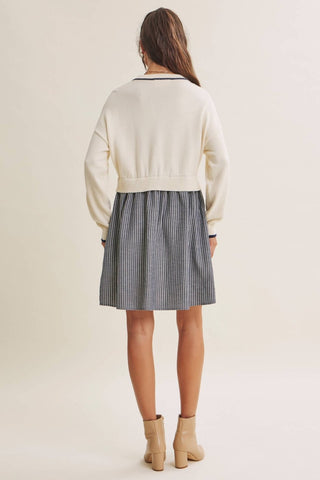 A woman viewed from behind wearing a cream knit cropped cardigan with dark trim over a gray and white vertically striped pleated skirt, paired with beige ankle boots, against a plain light beige studio background.