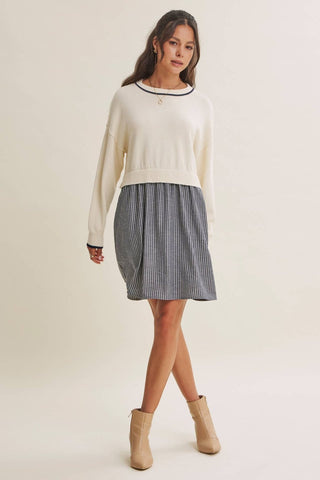 A woman modeling a cream knit sweater with a black crew neck trim, paired with a gray and white vertically striped pleated skirt and beige ankle boots, against a solid light beige studio background.