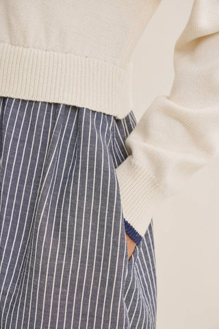 Close-up of a person wearing a cream ribbed knit sweater and navy-and-white vertically striped trousers, with a hand in the trouser pocket.