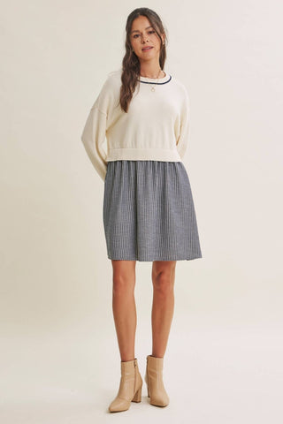 A woman modeling a cream knit crewneck sweater and a blue-and-white vertically striped pleated A-line skirt, paired with beige ankle boots, against a solid light beige studio background.