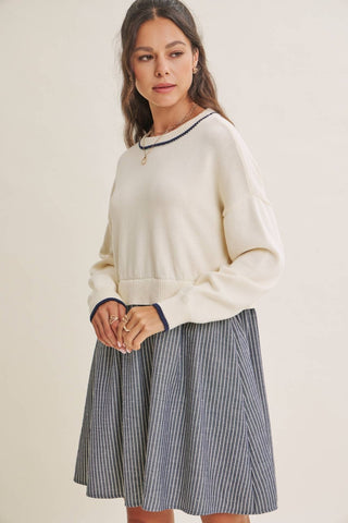 A woman modeling a cream knit sweater with dark navy trim and a navy and white vertically striped pleated skirt, set against a plain beige studio background.