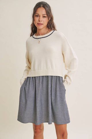 A woman modeling a cream knit crewneck sweater with black trim and a blue-and-white vertically striped pleated skirt, set against a plain beige studio background.