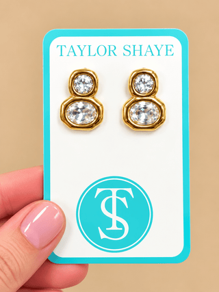 A close-up of a hand holding a jewelry display card featuring a pair of gold-toned stud earrings with clear faceted stones. The card is branded TAYLOR SHAYE with a turquoise logo and monogram, against a warm beige background.