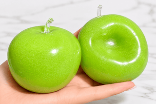 Two green apple-shaped squishes with clear stems are held in a person's hand against a light marble background.