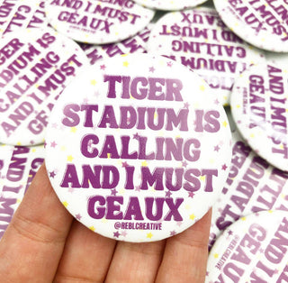 A hand holds a round white button badge with purple text reading 'TIGER STADIUM IS CALLING AND I MUST GEAUX' and small yellow star accents. Multiple similar badges are scattered in the background.