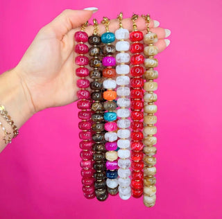 A hand holds six strands of beaded keychains against a bright pink background. The keychains feature round, polished beads in various colors including pink, brown, blue, white, orange, red, and beige, with gold-toned metal keychain hardware.