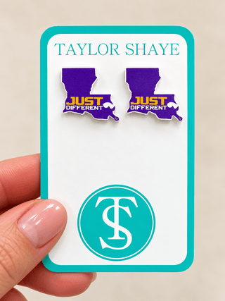 A hand holds a white display card featuring two purple Louisiana-shaped earrings with yellow and white 'JUST DIFFERENT' text. The card is branded 'TAYLOR SHAYE' with a teal circular 'TS' logo at the bottom.