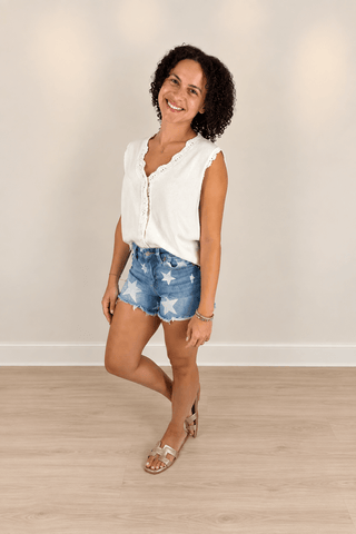 A woman modeling a white sleeveless top with scalloped edges and button details, paired with blue denim shorts featuring white star patterns, standing on a light wood floor against a neutral beige wall.
