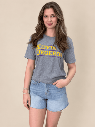 A woman modeling a heather gray graphic t-shirt with 'KIFFIN ORGERON' printed in yellow and purple across the chest, paired with light-wash denim shorts against a solid beige background.