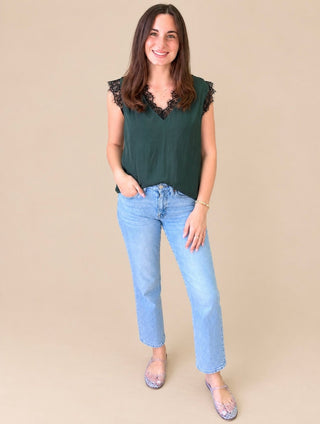 A woman modeling a dark green sleeveless top with black lace trim at the scoop neck and shoulders, paired with light blue jeans and patterned flats, against a solid beige background.