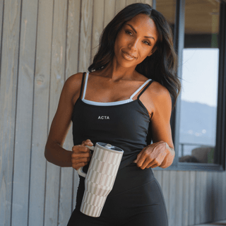 A woman wearing a black ACTA tank top and black leggings holds a striped ceramic tumbler. She is standing in front of a gray wood-paneled wall with a window in the background.