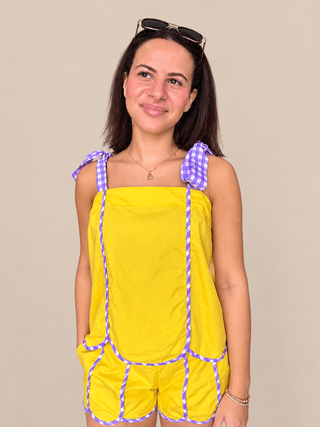 A woman wearing a bright yellow romper with purple and white gingham trim on the shoulder straps and along the seams, standing against a solid beige background.