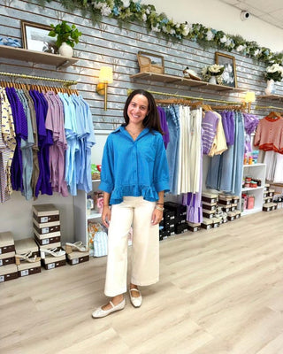 A woman stands in a clothing boutique wearing a bright blue ruffled button-up blouse and cream wide-leg pants. The background features a gray brick wall decorated with greenery, wooden shelves, and racks filled with colorful clothing.