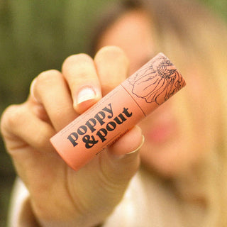 Hand holding a pink lip balm with 'poppy & pout' branding against a blurred background