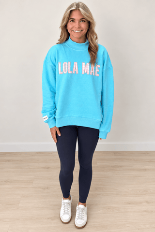 A woman modeling a bright turquoise blue mock neck sweatshirt with 'LOLA MAE' printed across the chest in pink and white, paired with dark navy leggings and white sneakers, standing against a neutral beige wall and light wood floor.