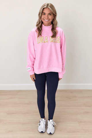 A woman modeling a pink sweatshirt with 'LOLA MIAE' printed in yellow and white across the chest, paired with dark leggings and white sneakers, standing against a neutral beige wall and light wood floor.