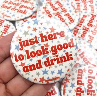 A hand holds a round pin-back button badge with the phrase 'just here to look good and drink' in red text over a white background with scattered red and blue stars. More identical badges are visible in the background.
