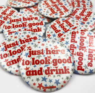 A pile of round pinback buttons featuring a white background with red and blue stars and the phrase 'just here to look good and drink' in red and blue lettering.