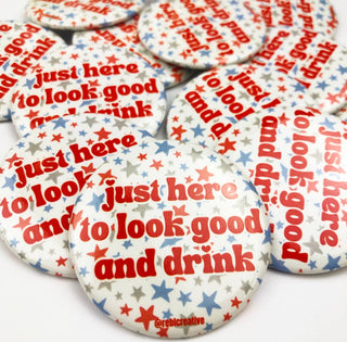 A pile of round pinback buttons featuring a patriotic red, white, and blue star pattern with the text 'just here to look good and drink' printed in red and blue.