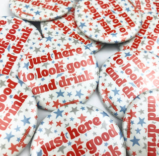 A pile of round pinback buttons featuring the phrase 'just here to look good and drink' in red text, surrounded by small red and blue stars on a white background.