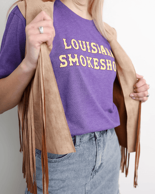Person wearing a purple tank top with 'Louisiana Smokesho' text, holding a beige fringed jacket.