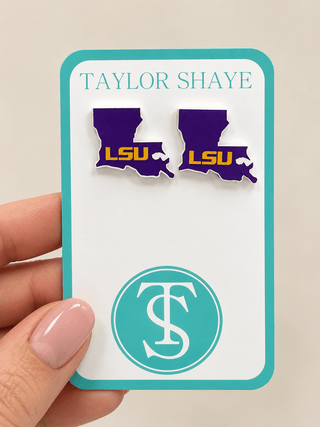 A hand holds a white display card featuring two purple Louisiana state-shaped pins with yellow LSU lettering, plus a teal circular monogram logo. The card reads TAYLOR SHAYE at the top.