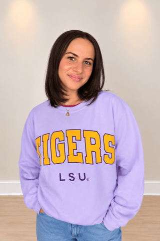 A woman with shoulder-length dark brown hair wearing a lavender purple LSU Tigers sweatshirt and blue jeans, standing against a neutral beige wall.