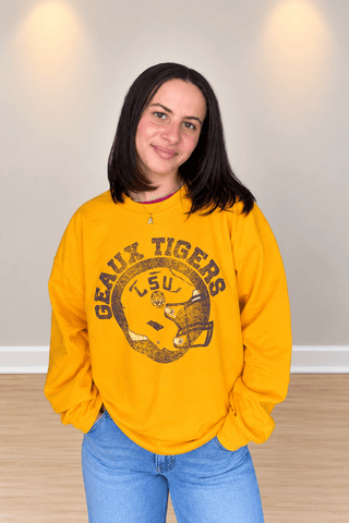 A young woman with dark hair smiling, wearing a bright yellow crewneck sweatshirt with a distressed 'Geaux Tigers' graphic on the front, paired with blue jeans, standing against a neutral beige wall.
