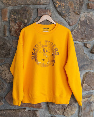 A bright yellow crewneck sweatshirt with a distressed brown 'Geaux Tigers LSU' graphic on the front, hanging on a wooden hanger against a rustic stone wall.