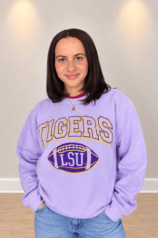 A woman wearing a lavender purple LSU Tigers sweatshirt with a football graphic, paired with blue jeans, standing against a neutral indoor wall.
