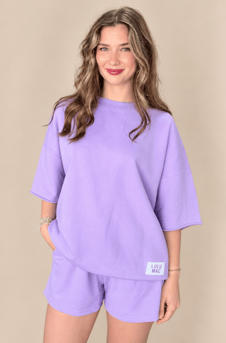 A woman modeling a matching lavender lounge set consisting of an oversized short-sleeve top and shorts, featuring a small white brand label on the bottom hem, set against a solid beige background.