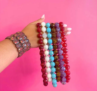 A hand with white manicured nails holds five strands of faceted round beads in red, white, light blue, brown, and purple, while wearing a chunky pink rhinestone bracelet against a solid pink background.