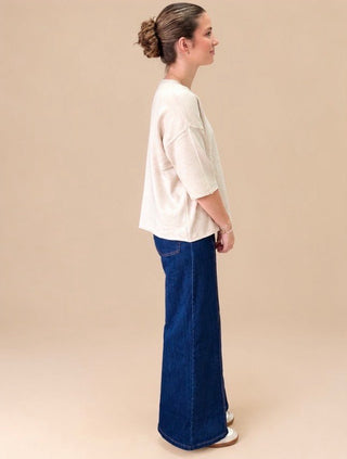 A woman shown in profile wearing a cream-colored short-sleeve top and dark blue wide-leg denim jeans, standing against a solid beige studio background.