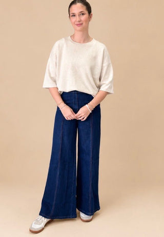 A woman modeling a cream-colored short-sleeve knit top and dark blue wide-leg denim pants against a solid beige studio background.