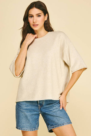A woman modeling an oversized short-sleeve t-shirt in a light oatmeal beige color, paired with blue denim shorts, against a solid light beige background.