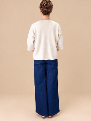 A woman viewed from behind wearing a cream-colored knit top and dark blue wide-leg jeans, standing against a solid beige background.