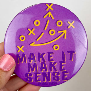 A round purple button badge held by a hand, featuring a yellow tactical crossword-style diagram and the text 'MAKE IT MAKE SENSE' in raised purple letters with yellow outlines.