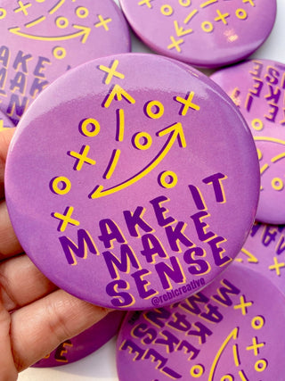 A close-up of a hand holding a round purple pinback button with yellow tactical football play graphics and the text 'MAKE IT MAKE SENSE' and '@rebelcreative'. More matching purple buttons are visible in the background.