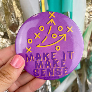 A hand with pink nail polish holds a round purple button badge featuring a yellow football play diagram and the text 'MAKE IT MAKE SENSE'. The background is a colorful, out-of-focus setting with metallic and teal elements.