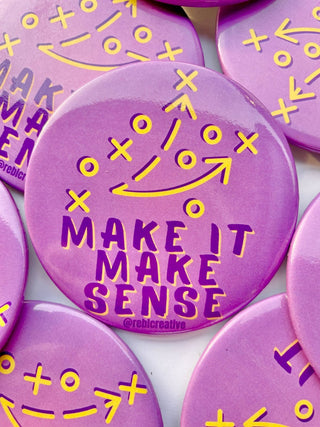 A close-up of round purple buttons with yellow tactical play symbols and the phrase “MAKE IT MAKE SENSE” printed on them.