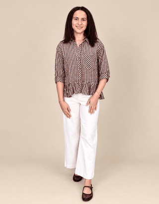 A woman modeling a brown and white checkered button-down shirt with a ruffled hem, paired with white wide-leg pants and dark brown shoes, against a solid beige background.